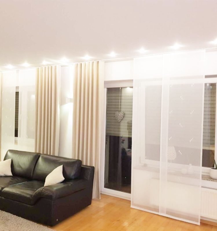 Wohnbereich mit großen Fensterfronten und transparenten Gardinen von M-Gardindendesign für eleganten Sicht- und Lichtschutz am Abend.